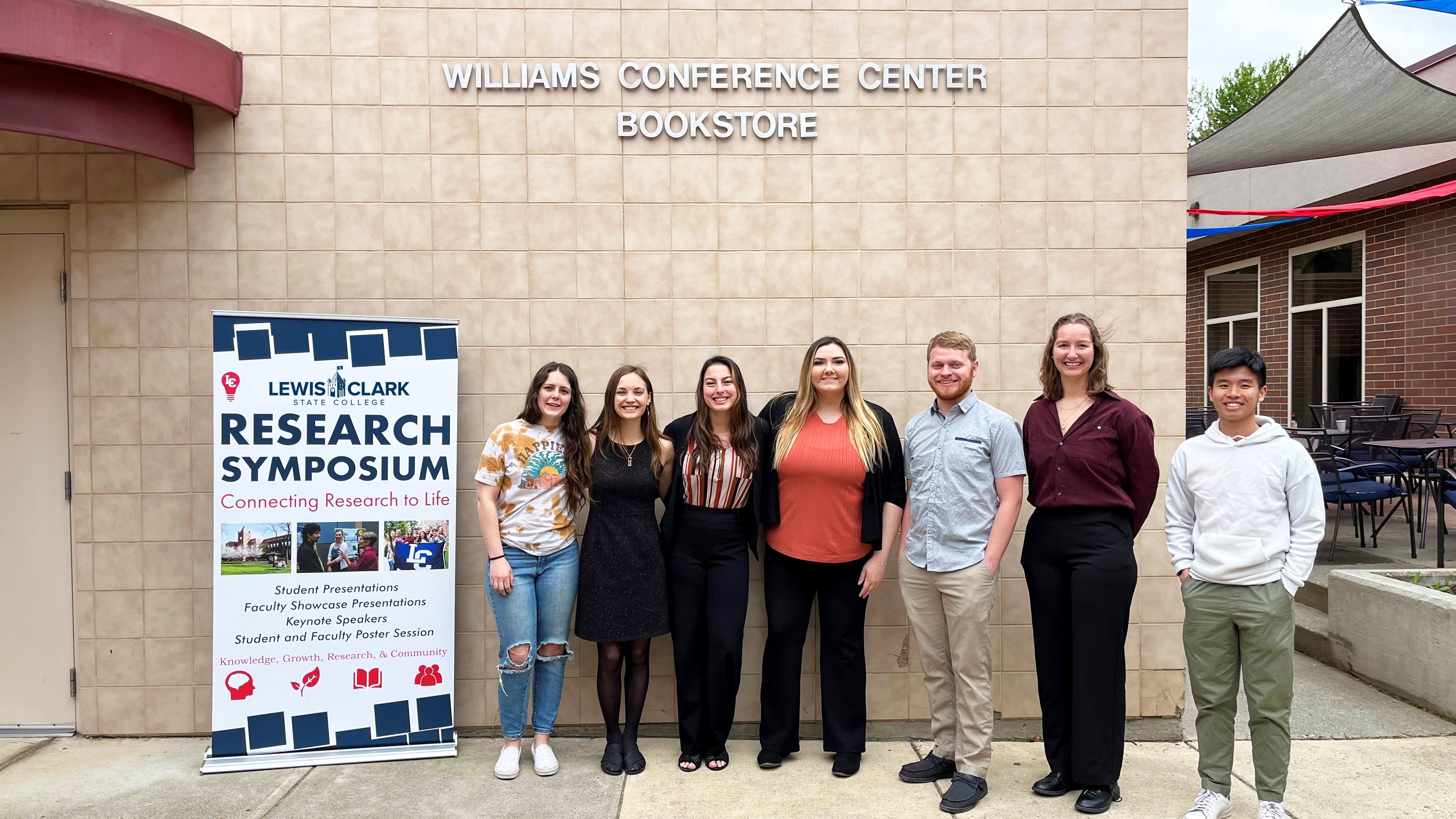 Seven people posed outside of William Conference Center by Research Symposium Sign