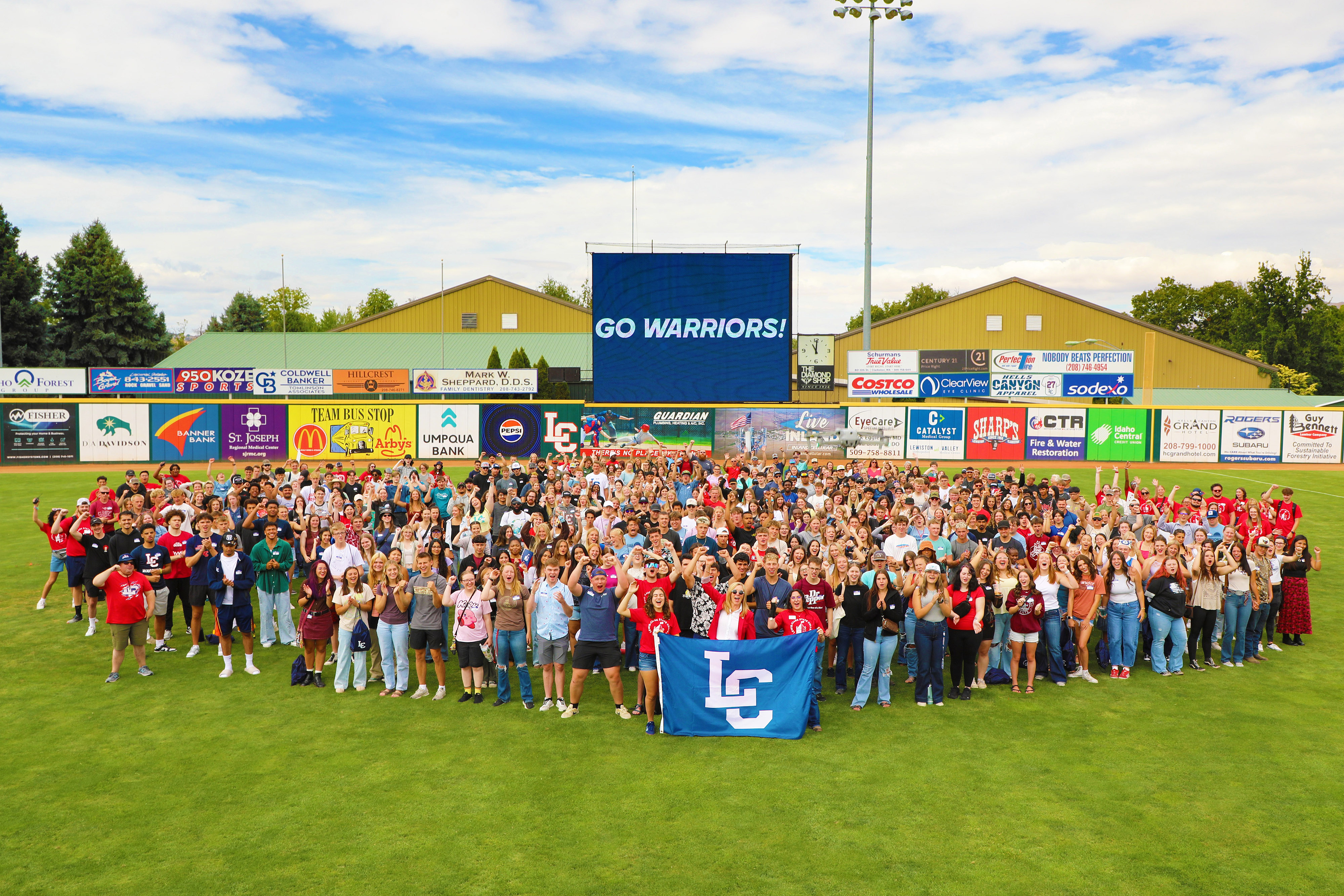 Group of students and LC State President holding up LC Flag cheering "Go Warriors" with sign in background saying "GO WARRIORS"
