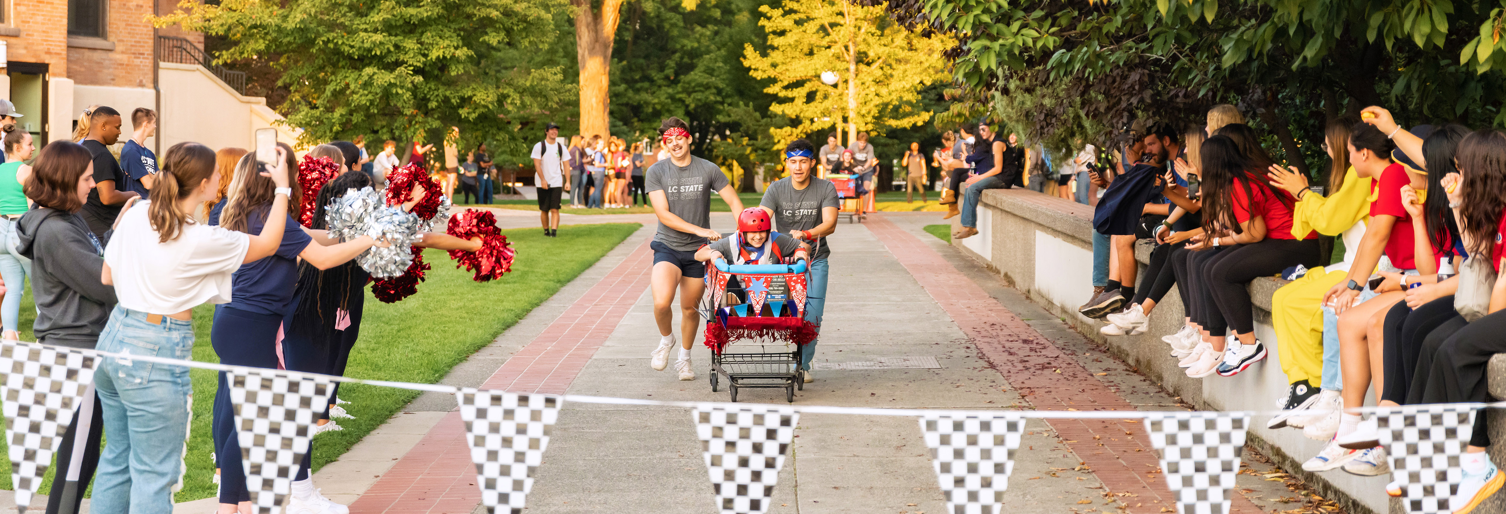 Two students push another student in a shopping cart as they prepare to cross the finish line with other students cheering them on
