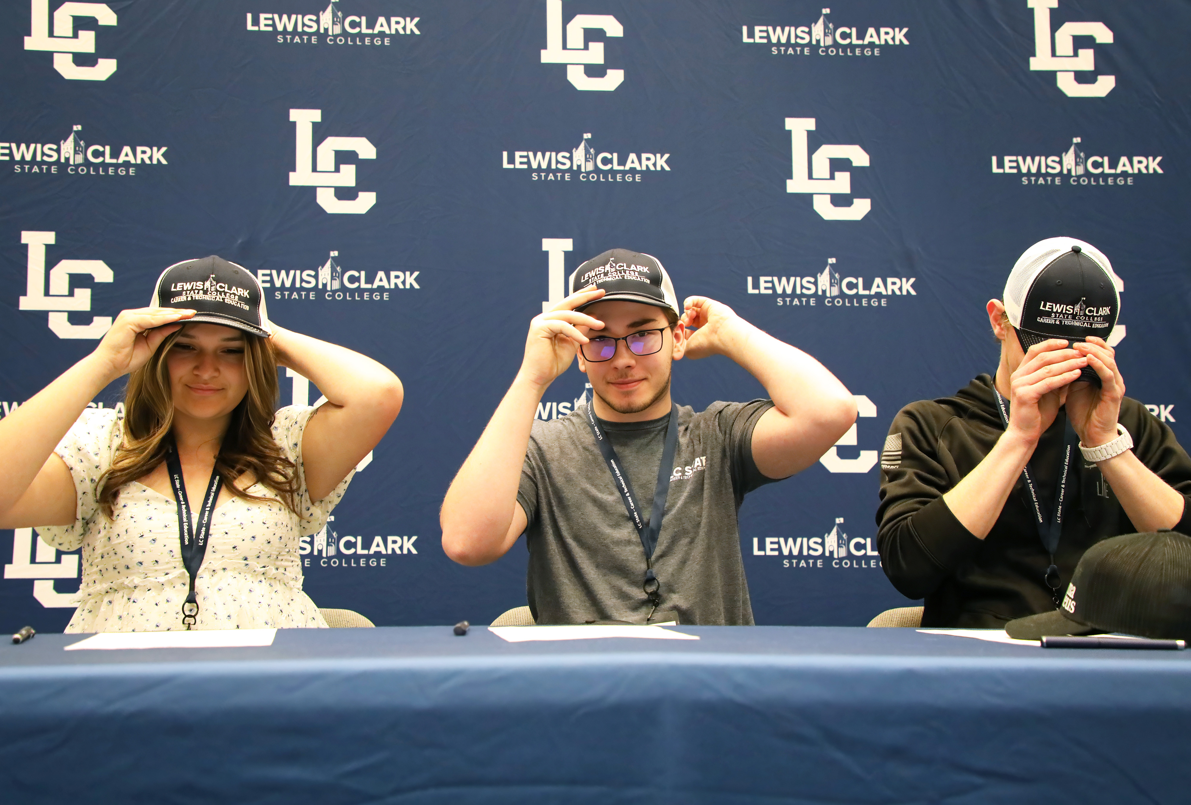 Three students put on Lewis-Clark State College hats in front of LC State backdrop