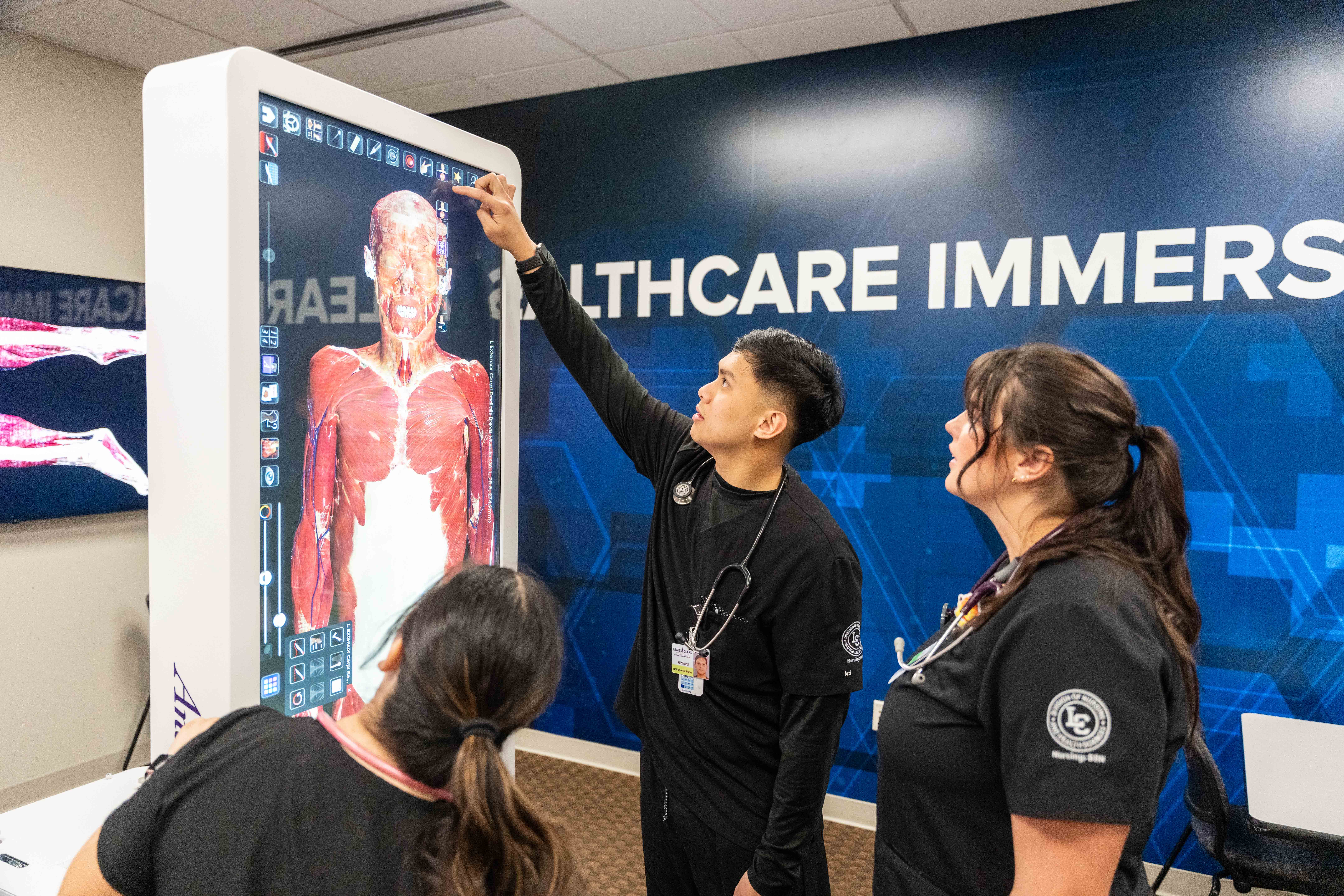 Three students in black scrubs examine a digital anatomy model on a large touchscreen in Healthcare Immersive Learning Lab