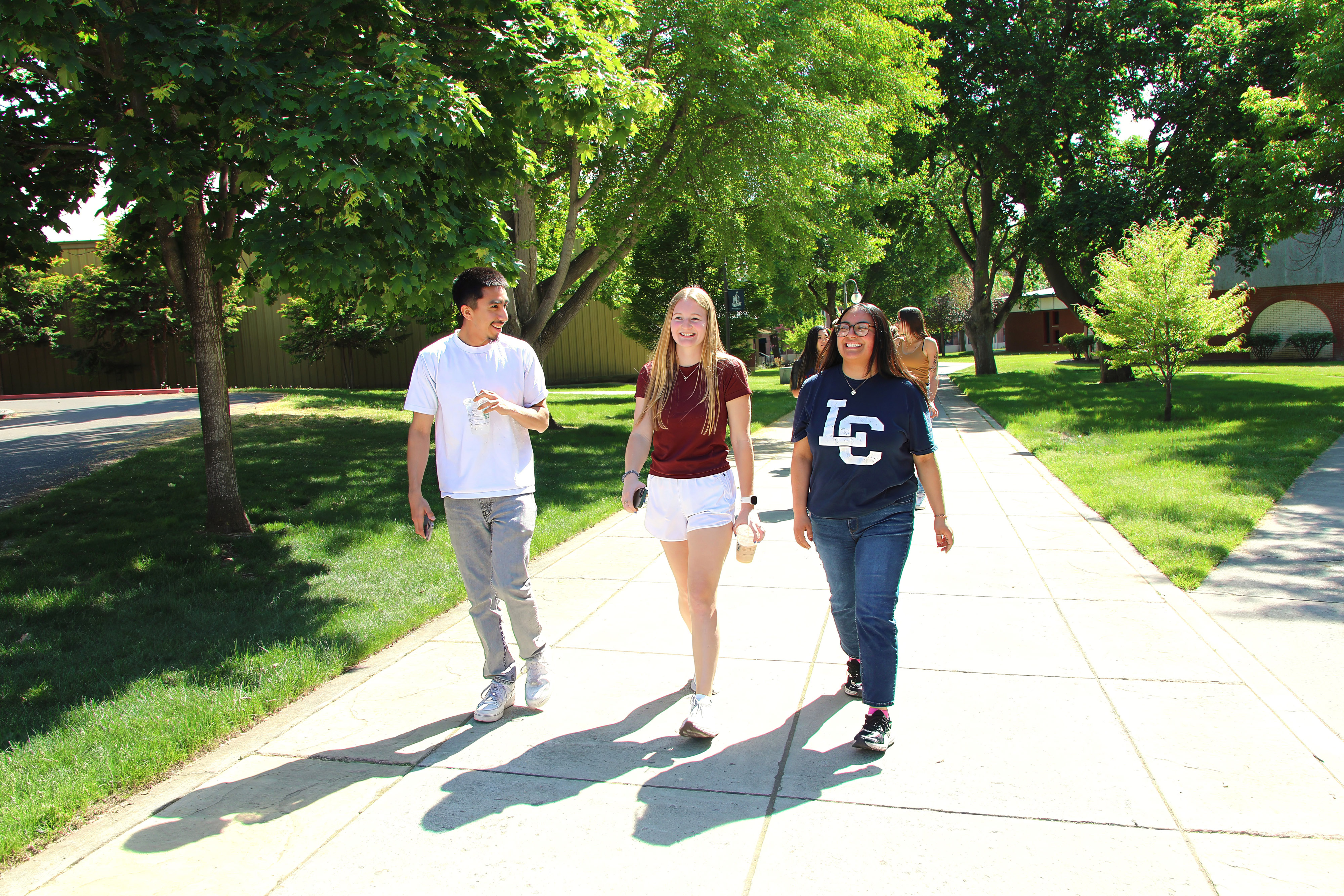 Three students walking and talking on sidewalk pathway on campus with two students behind them and buildings in distance